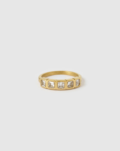 A gold band ring featuring a row of six square-cut clear gemstones in a bezel setting, shown against a solid light gray background.