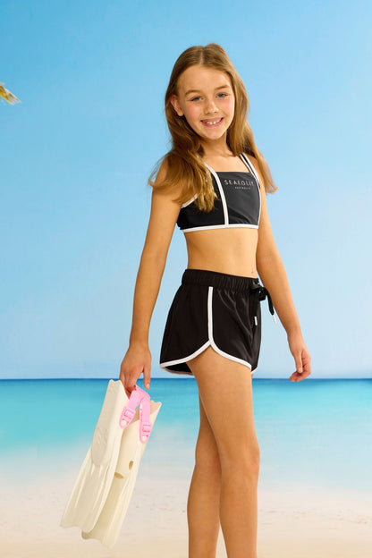 Seafolly Girls Essentials Boardshort in Black features an elasticated waistband, draw cord, scallop hemline, contrast piping.